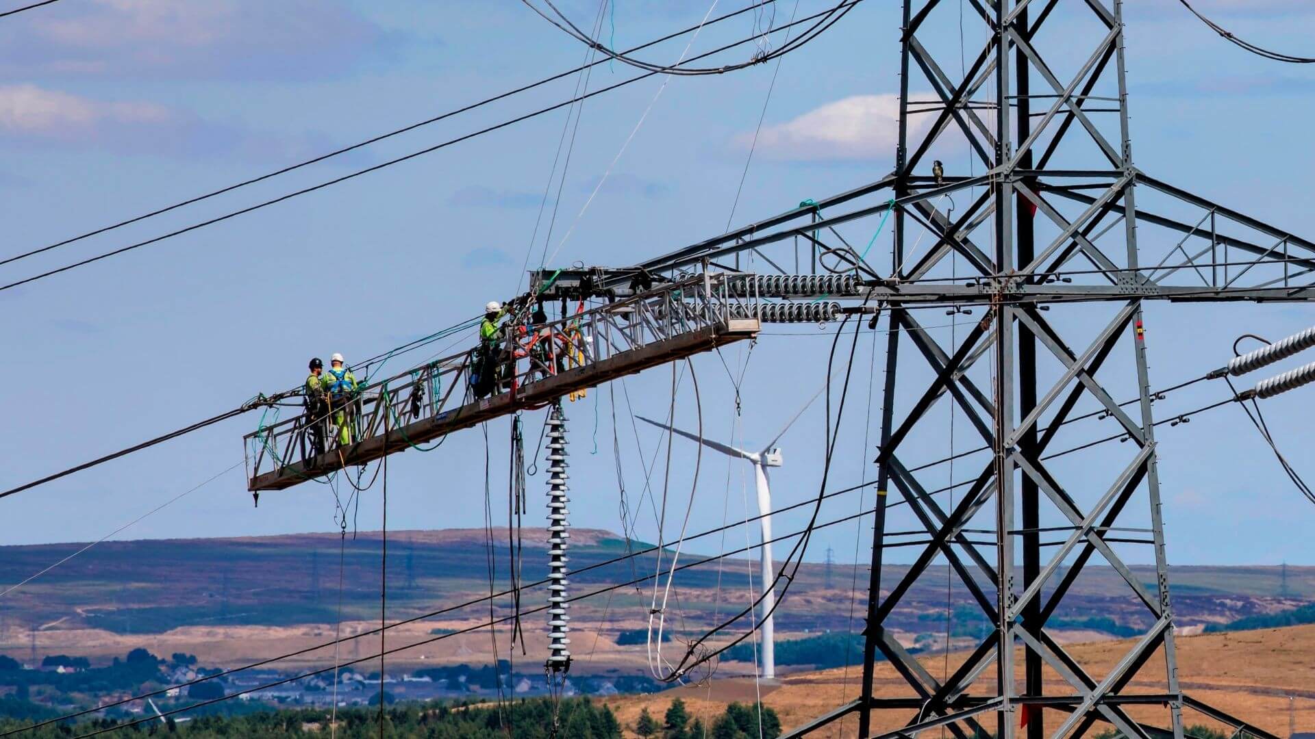 Three engineers in high-viz clothing and hard hats, working to install new equipment from a gantry suspended under electricity transmission line connection to pylon