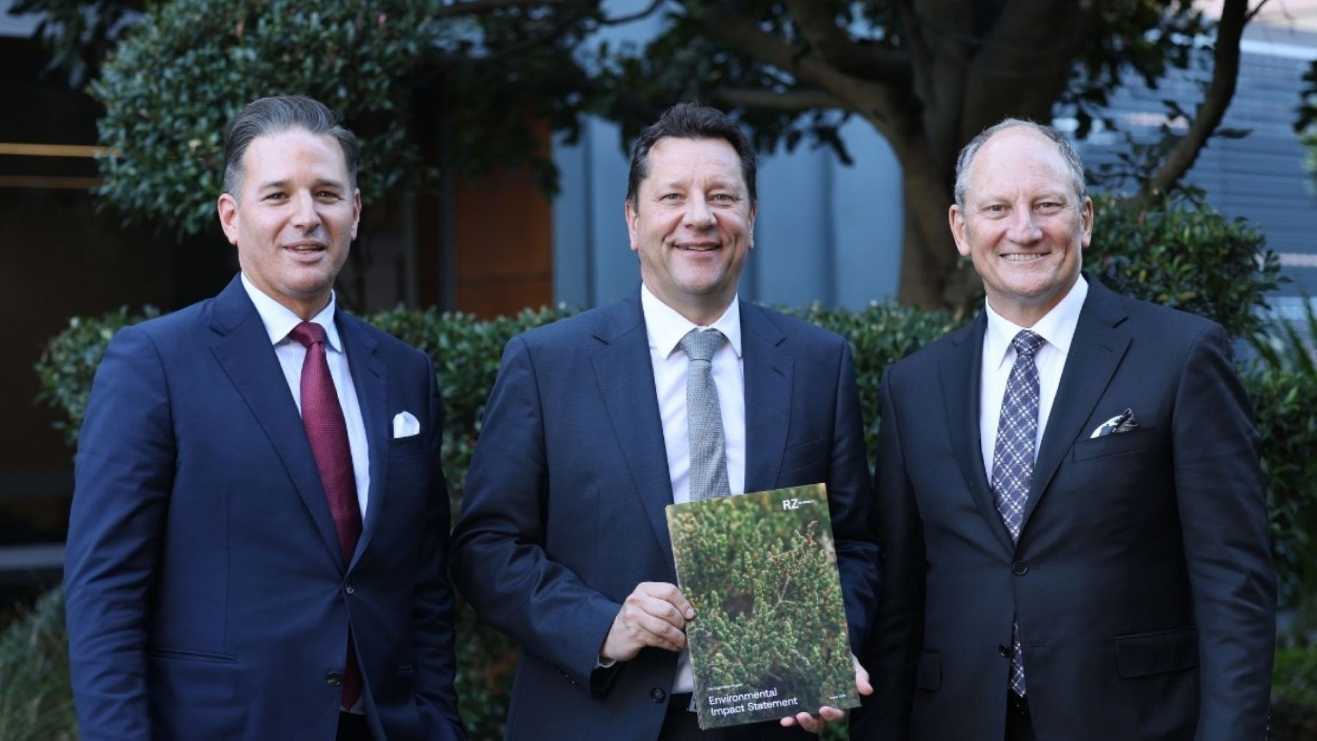 Three men in suits, stood side by side outside and smiling to camera; man in centre holding a printed brochure
