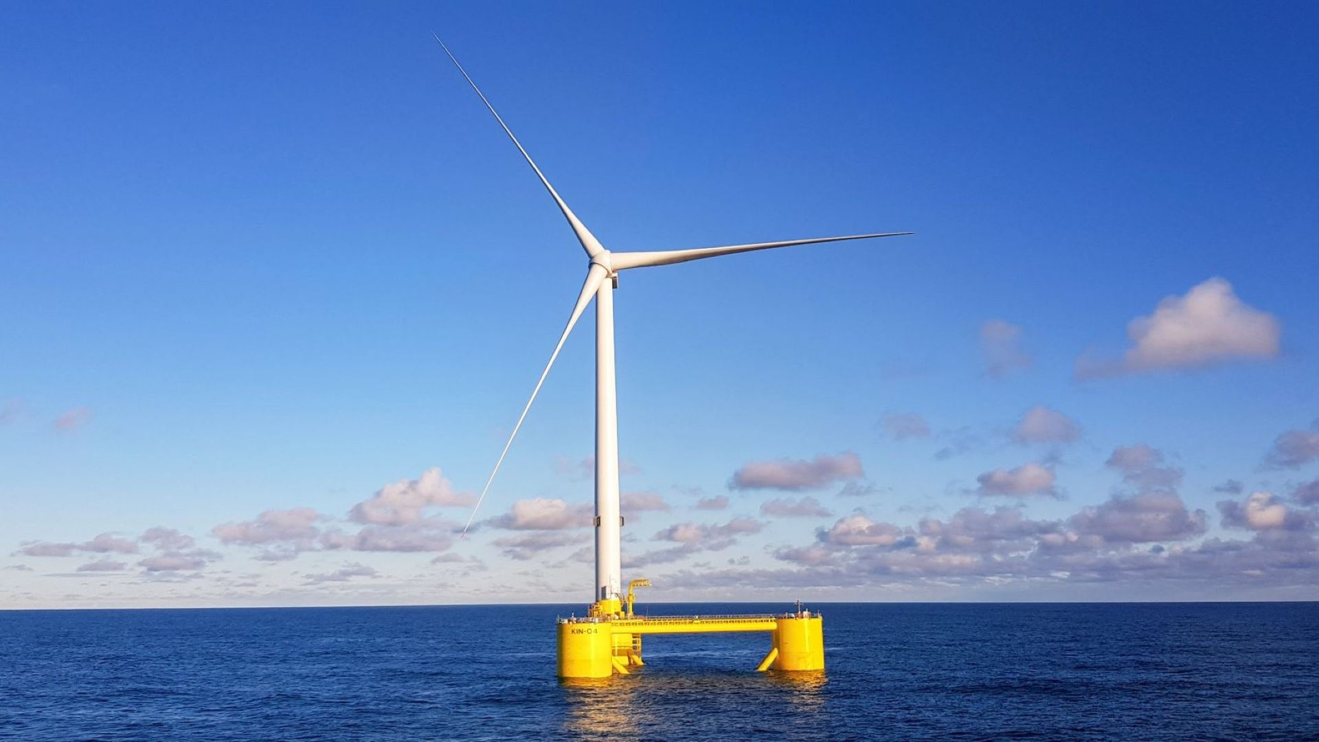 Three-bladed wind turbine on yellow floating foundations, in calm blue sea, with blue sky and fluffy white clouds in background