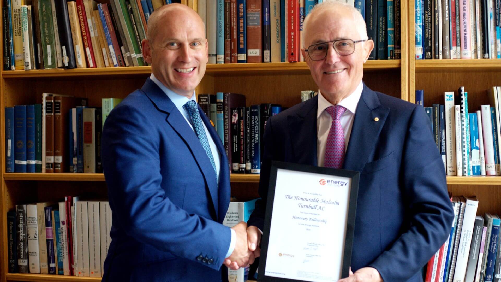 Two men standing side by side, shaking hands and smiling to camera, the one on the right holding a framed certificate. They are standing in front of library bookshelves filled with books.
