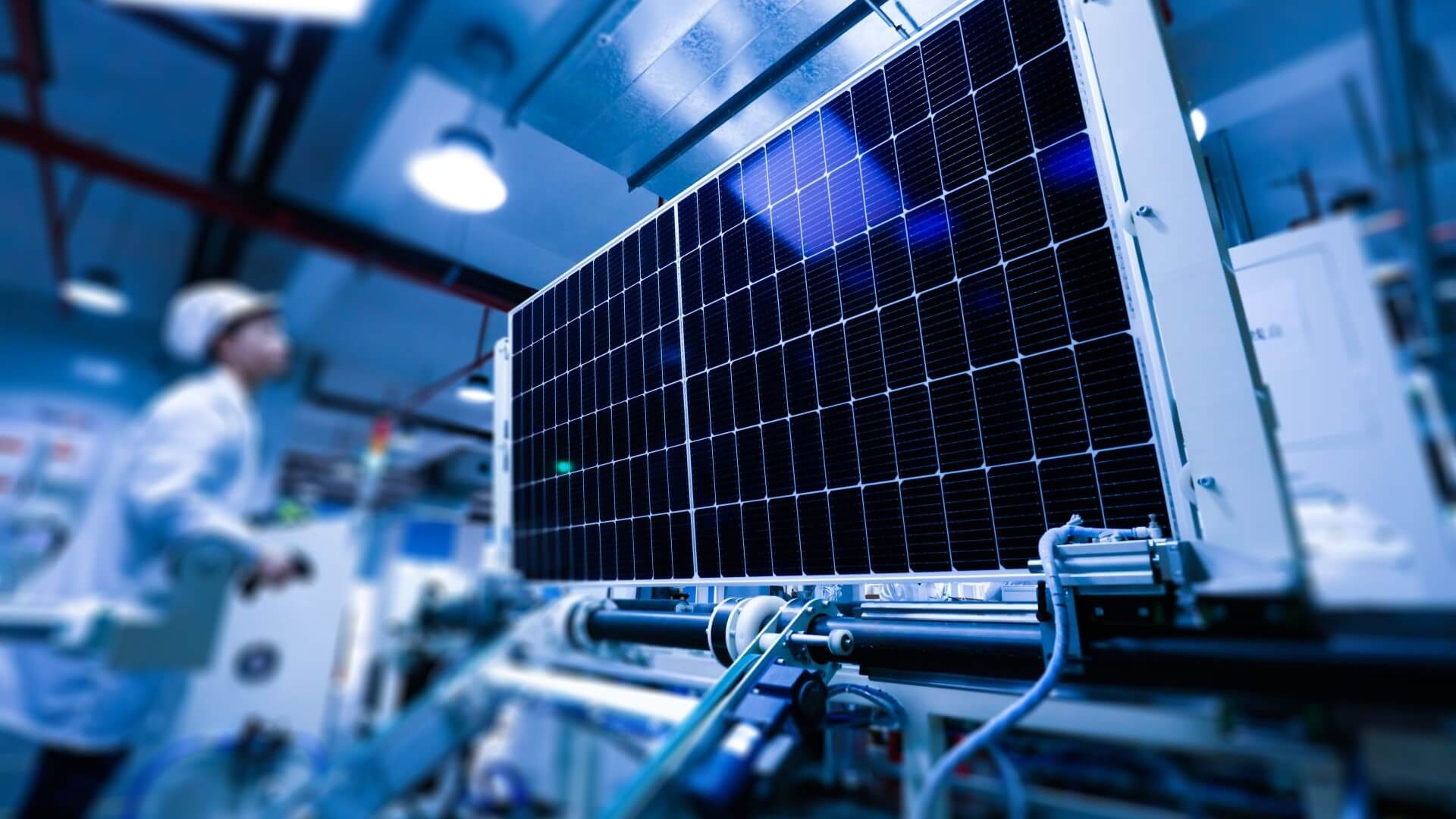 Close up of a solar panel standing upright on a manufacturing factory conveyor, with a worker visible in the background of the photo