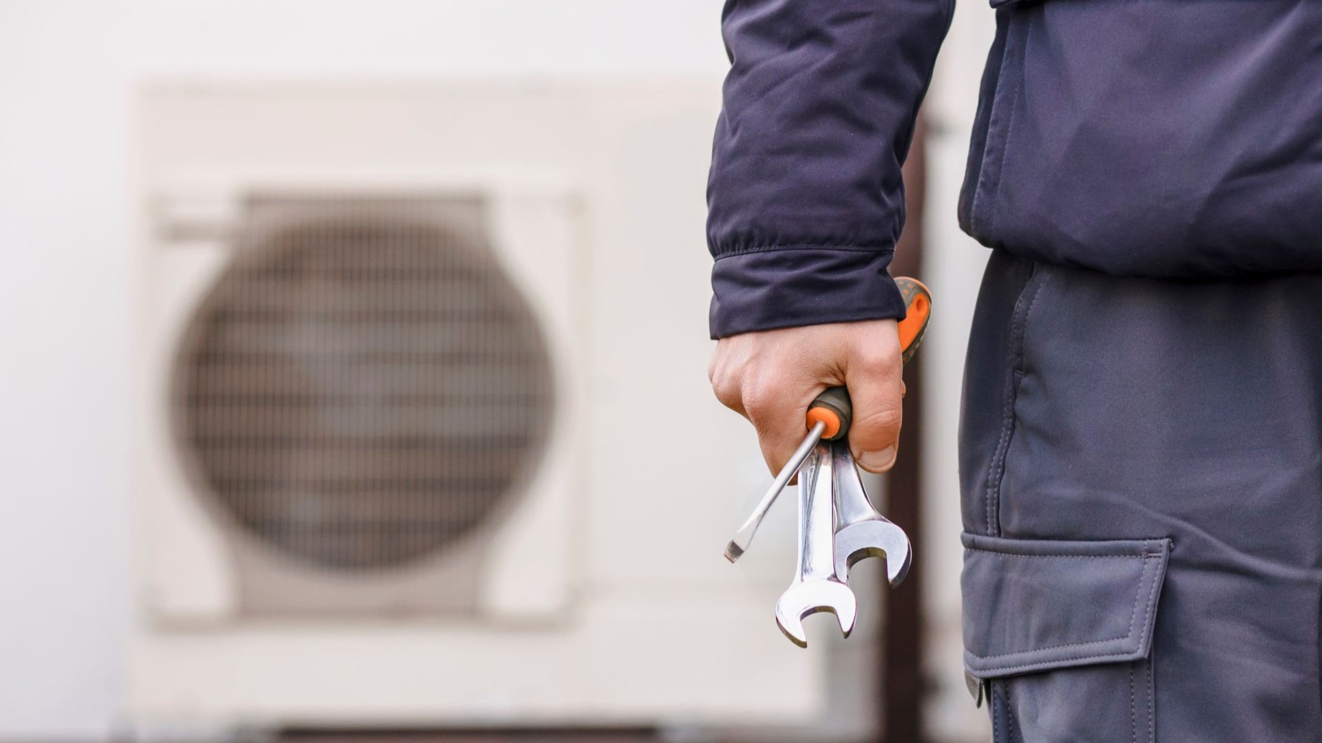 Close up of the right wrist of an engineer in a dark blue boiler suit, holding a screwdriver and two wrenches in hand, walking away from a blurred heat pump that is visible in the background.