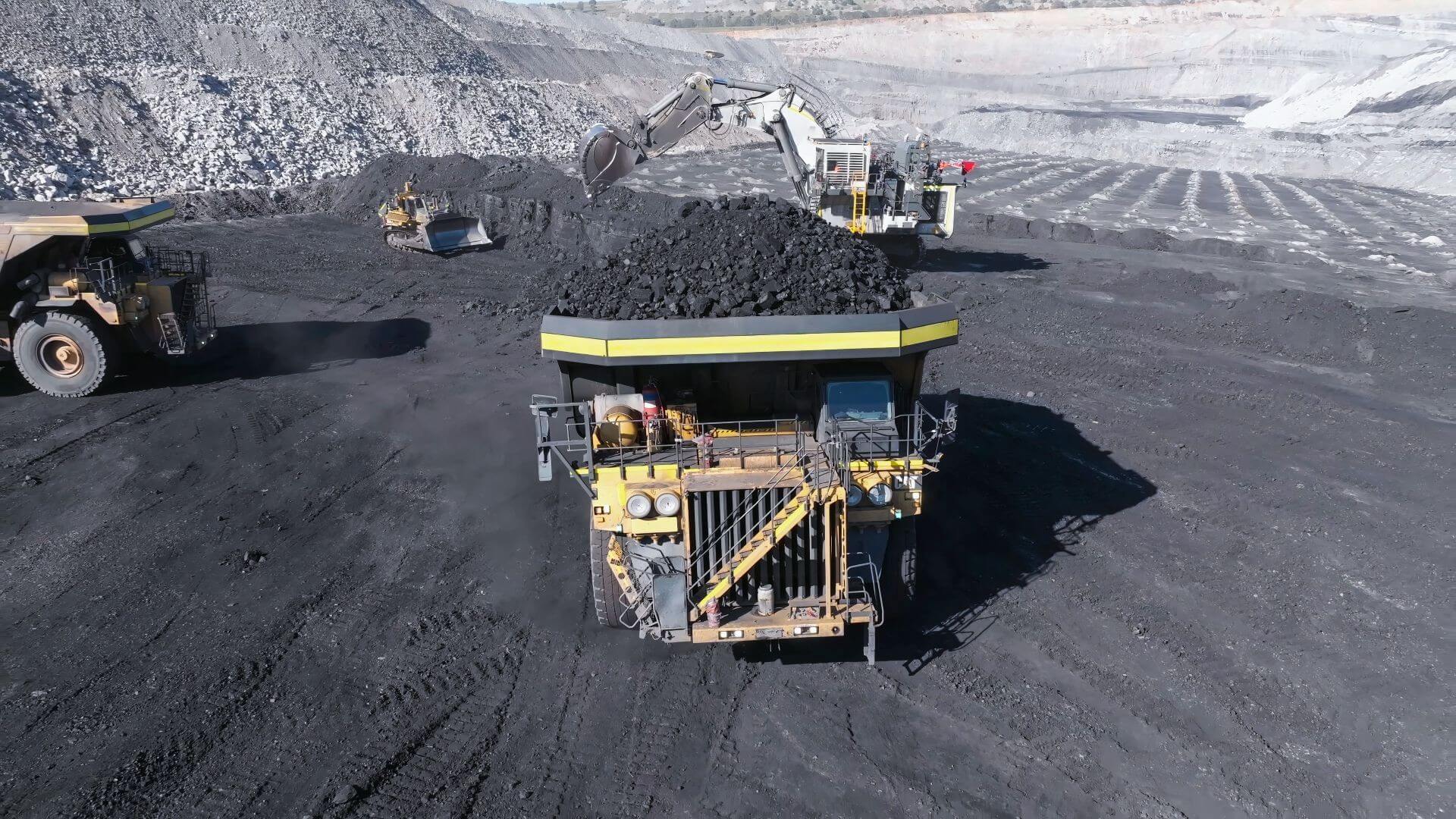 Operations in an open cast mine with very large truck loaded with coal in foreground of picture with another to the side and a digger in the background