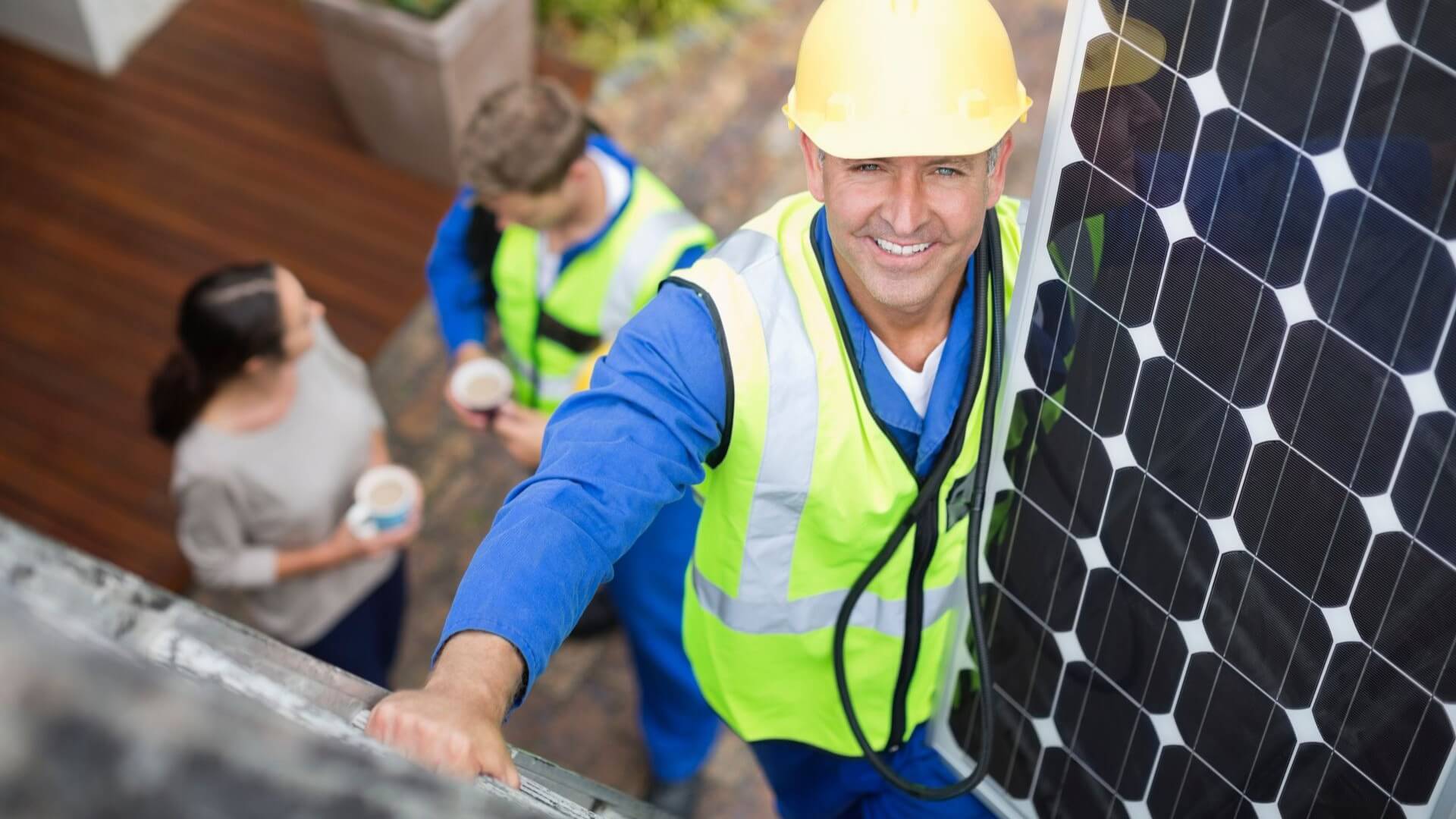 Man in high viz jacket and hard hat, standing on scaffold at height next to a solar panel, and smiling to camera, with two people standing at ground level, talking