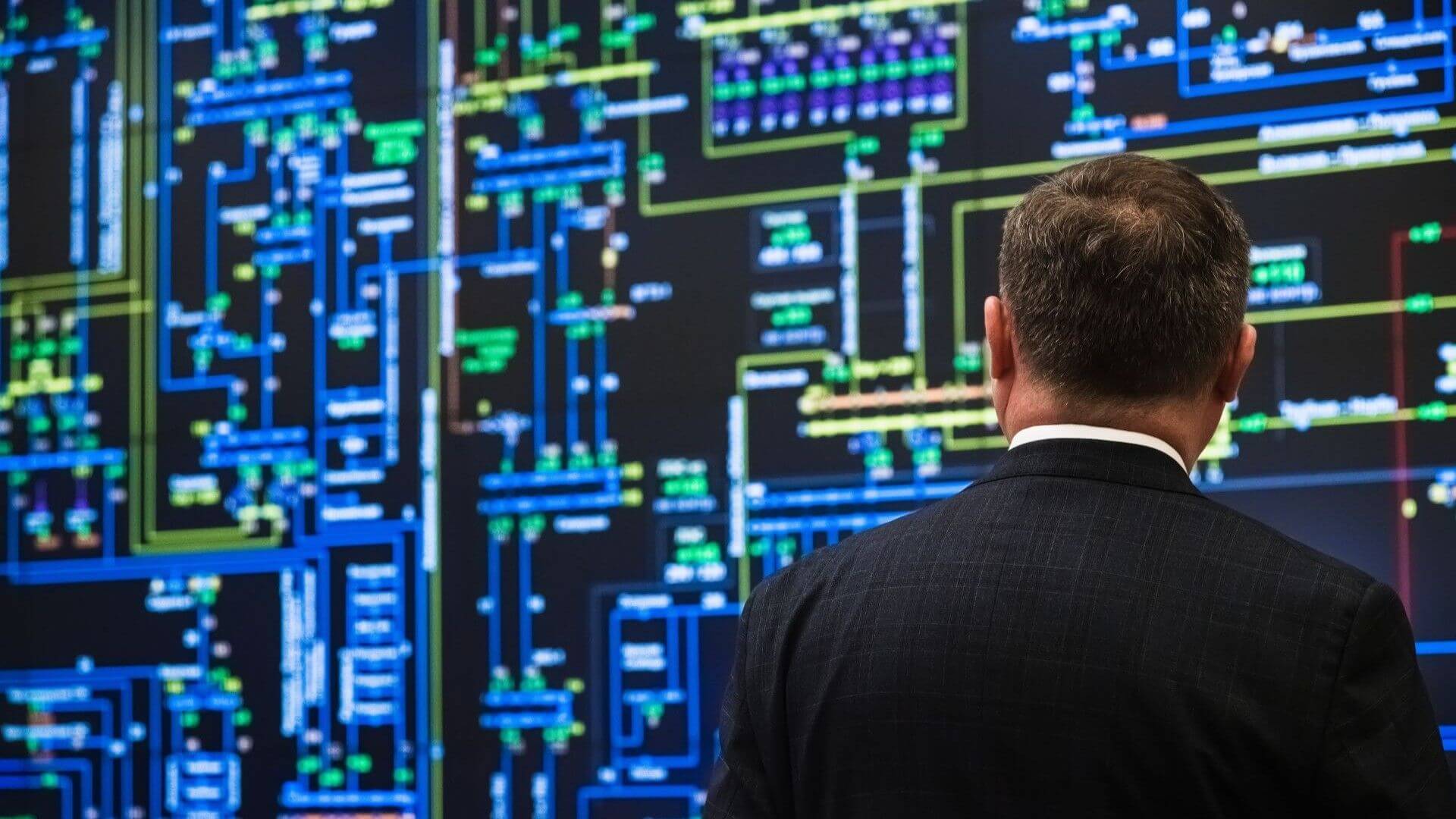 Man standing in front of large computer screen showing lots of data points, switches and connections in blues, yellows and green colours