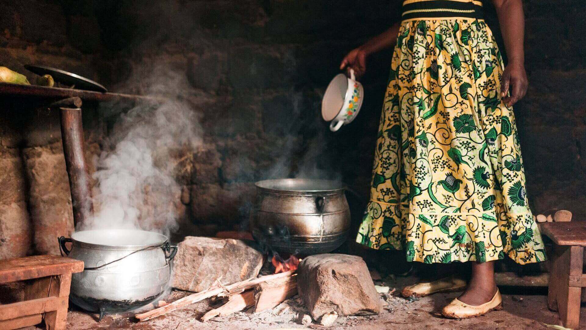 Close up of lower half of a woman in patterned dress standing beside metal two metal cooking pots over wood fire on floor inside a rural hut