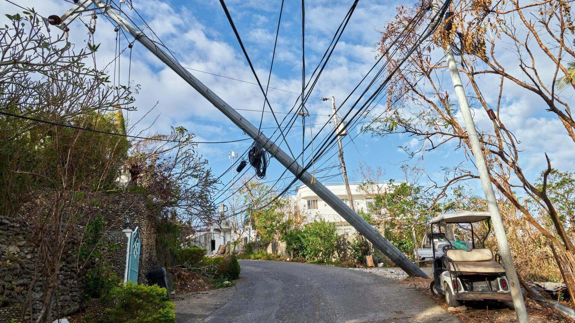 Typhoon damage: mast holding power lines fallen on to telephone lines, with battered golf cart style buggy to right, next to damaged trees and fence, and building in background