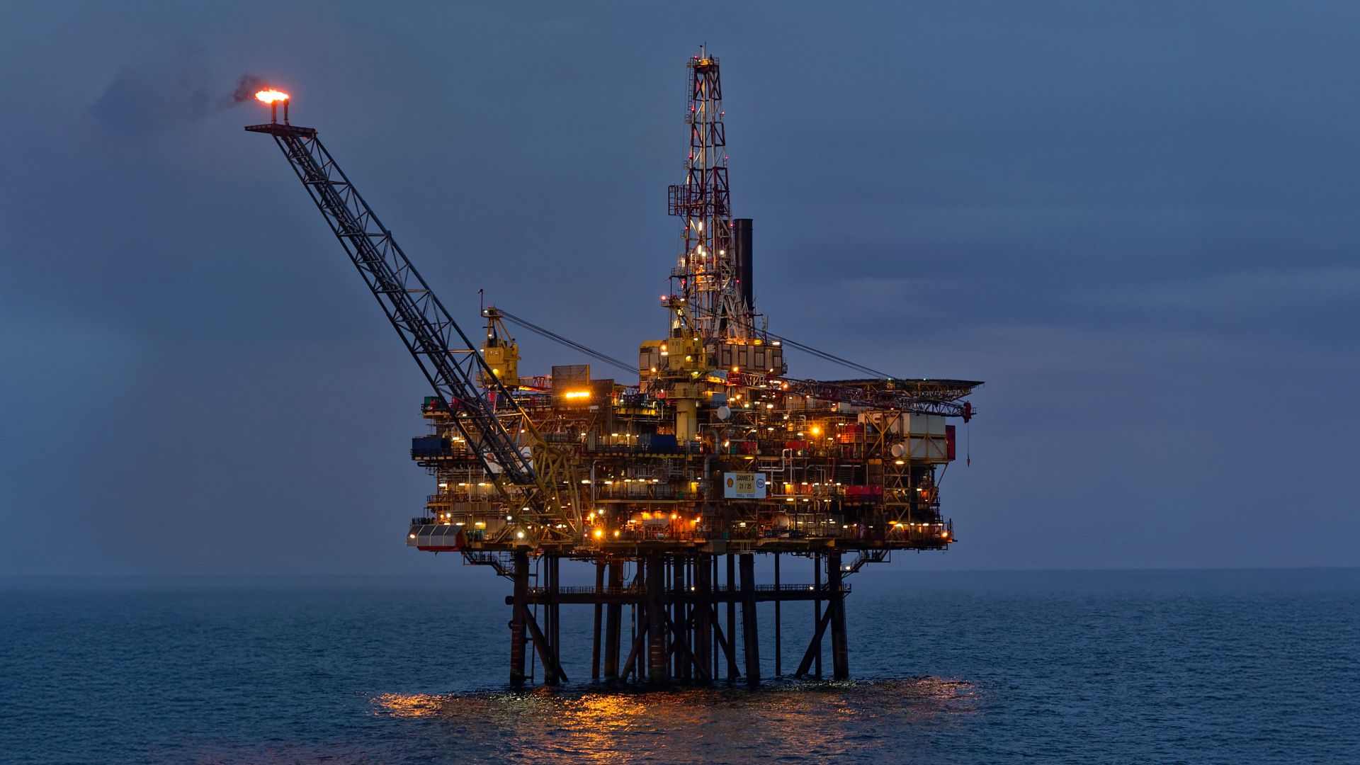 Offshore platform at night, showing bright orange of gas flare and other installation lighting, reflected in calm sea