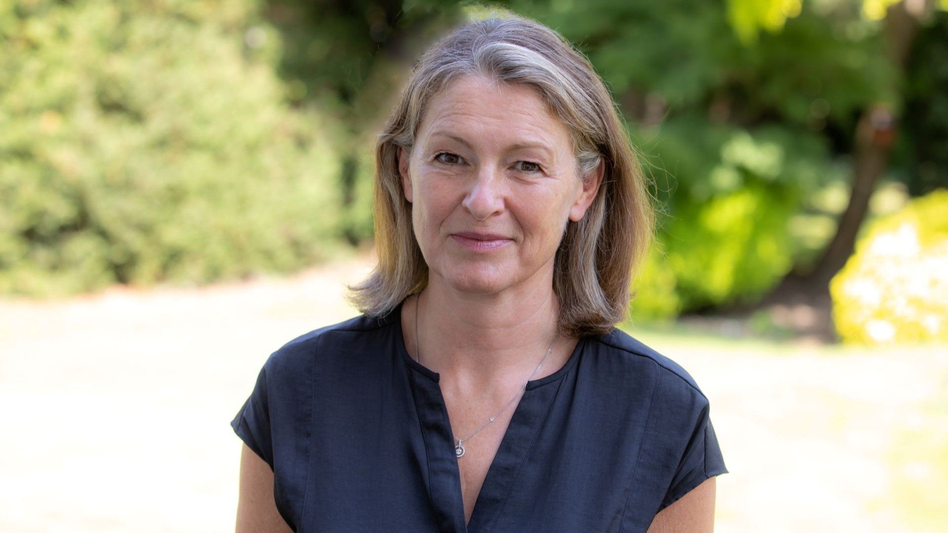 Head and shoulders photo of Anne Murrell, standing outside with green foliage blurred in the background
