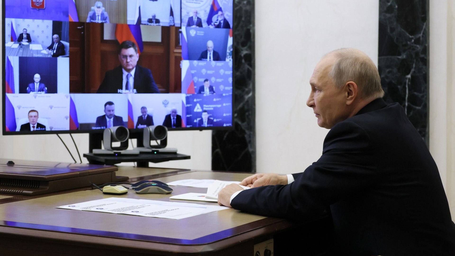 Russian President Vladimir Putin, sat, with hands resting on a desk, in front of a computer monitor showing videoconference participants